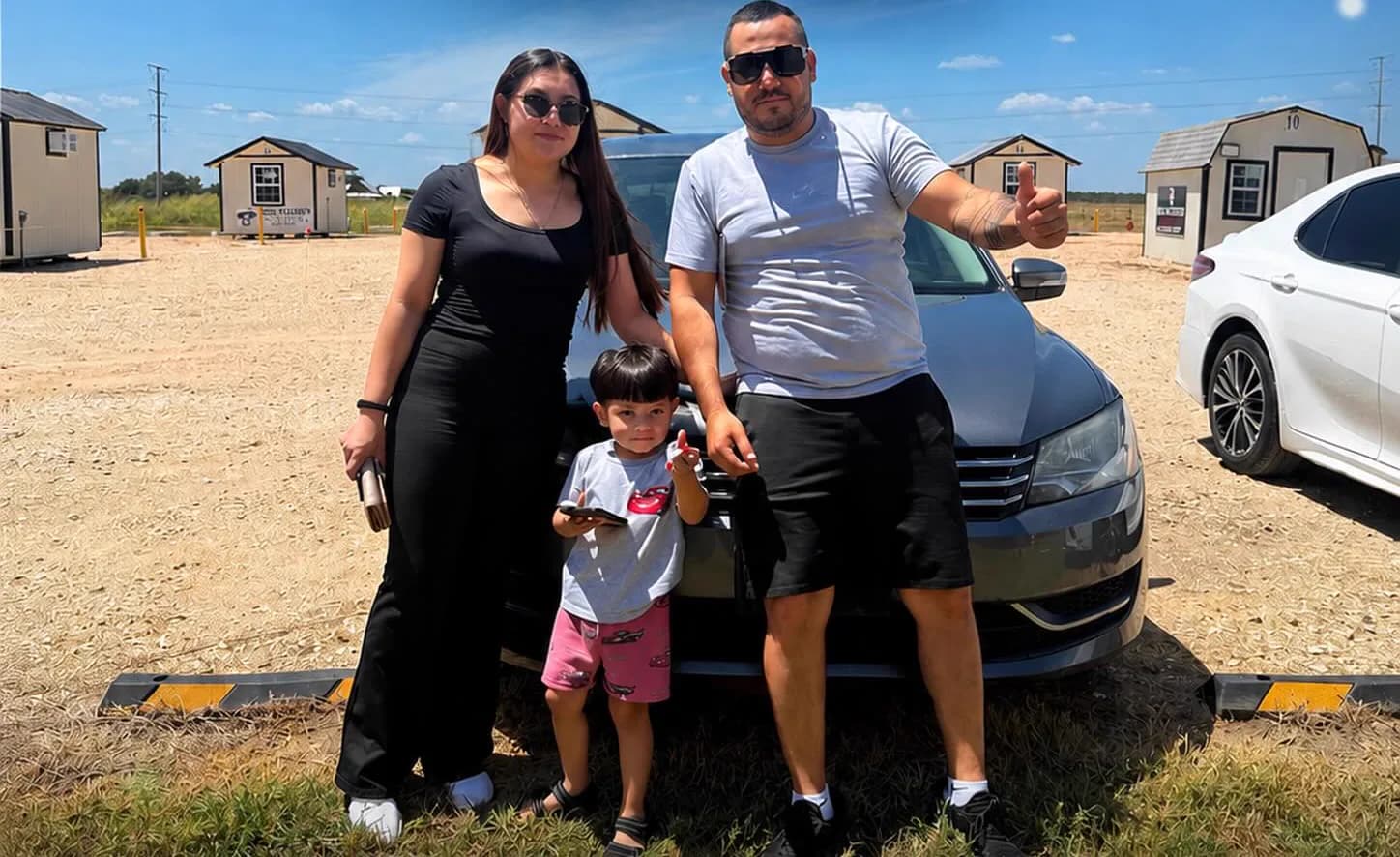 A family of three standing in front of the grey Volkswagen Passat they bought, on the gravel lot in Hutto.